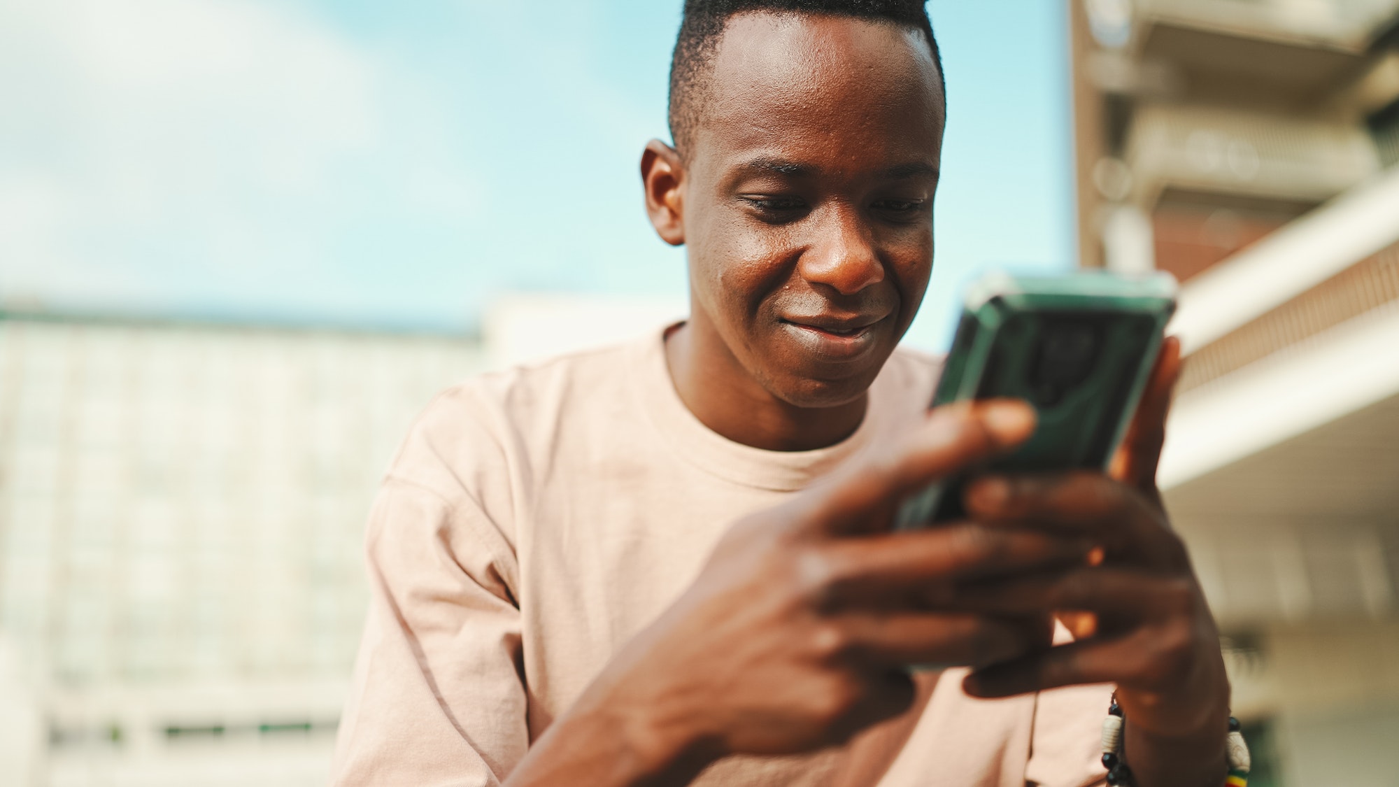 Young african student sits outside of university, uses cellphone, taps on smartphone screen